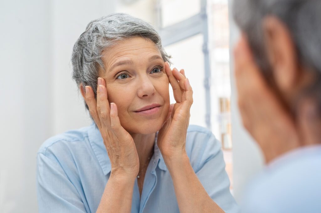 Mature woman examining her eyes in the mirror, considering eyelid surgery (blepharoplasty) for facial rejuvenation. Dr. Deuber in Dallas, TX
