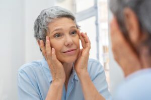 Mature woman examining her eyes in the mirror, considering eyelid surgery (blepharoplasty) for facial rejuvenation. Dr. Deuber in Dallas, TX